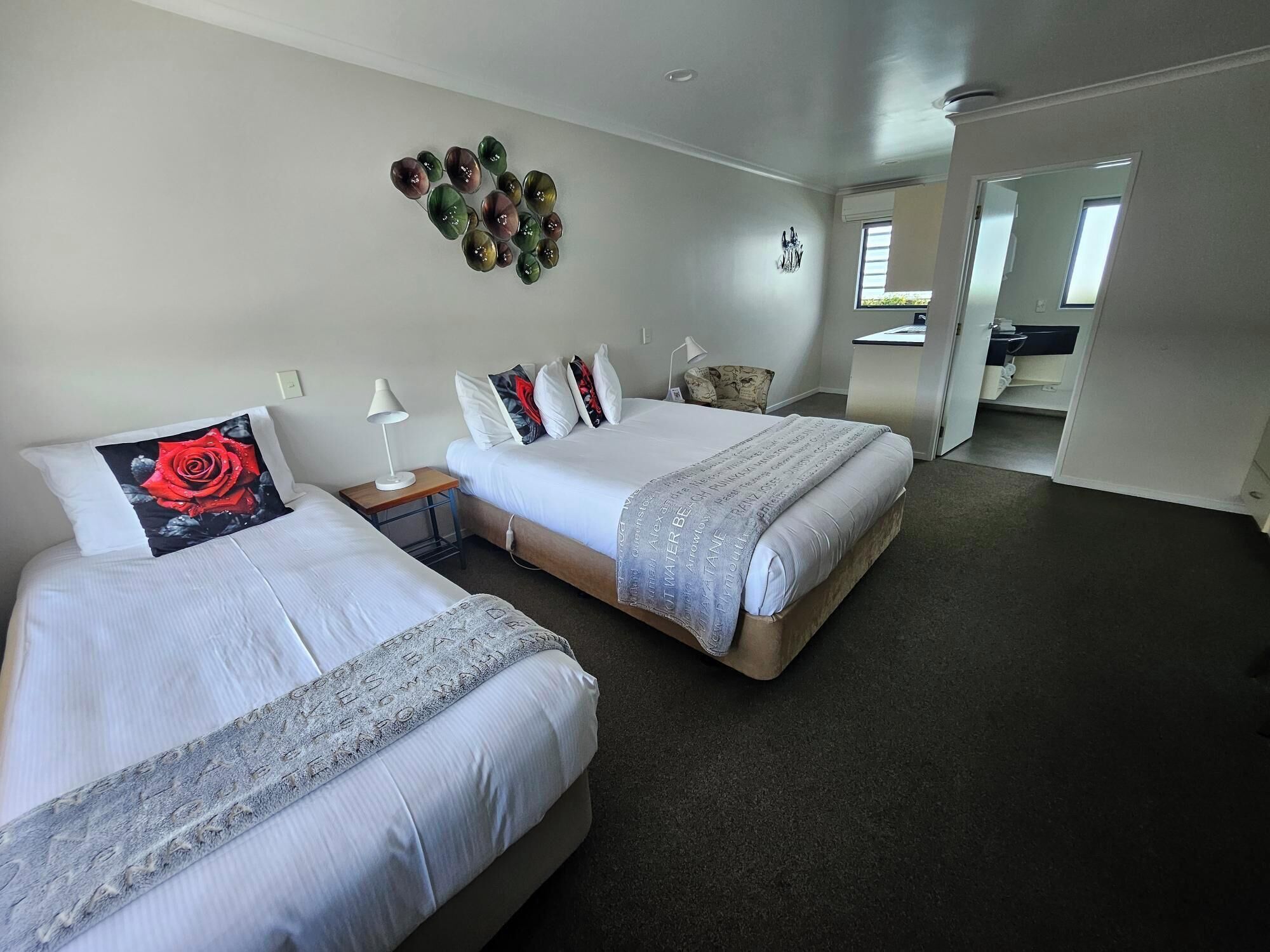 A hotel room with two beds, a small table, a lamp, a decorative wall piece, and a view of the bathroom and kitchen through an open door.
