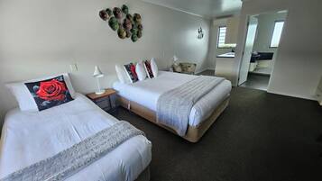 A hotel room with two beds, a small table, a lamp, a decorative wall piece, and a view of the bathroom and kitchen through an open door.