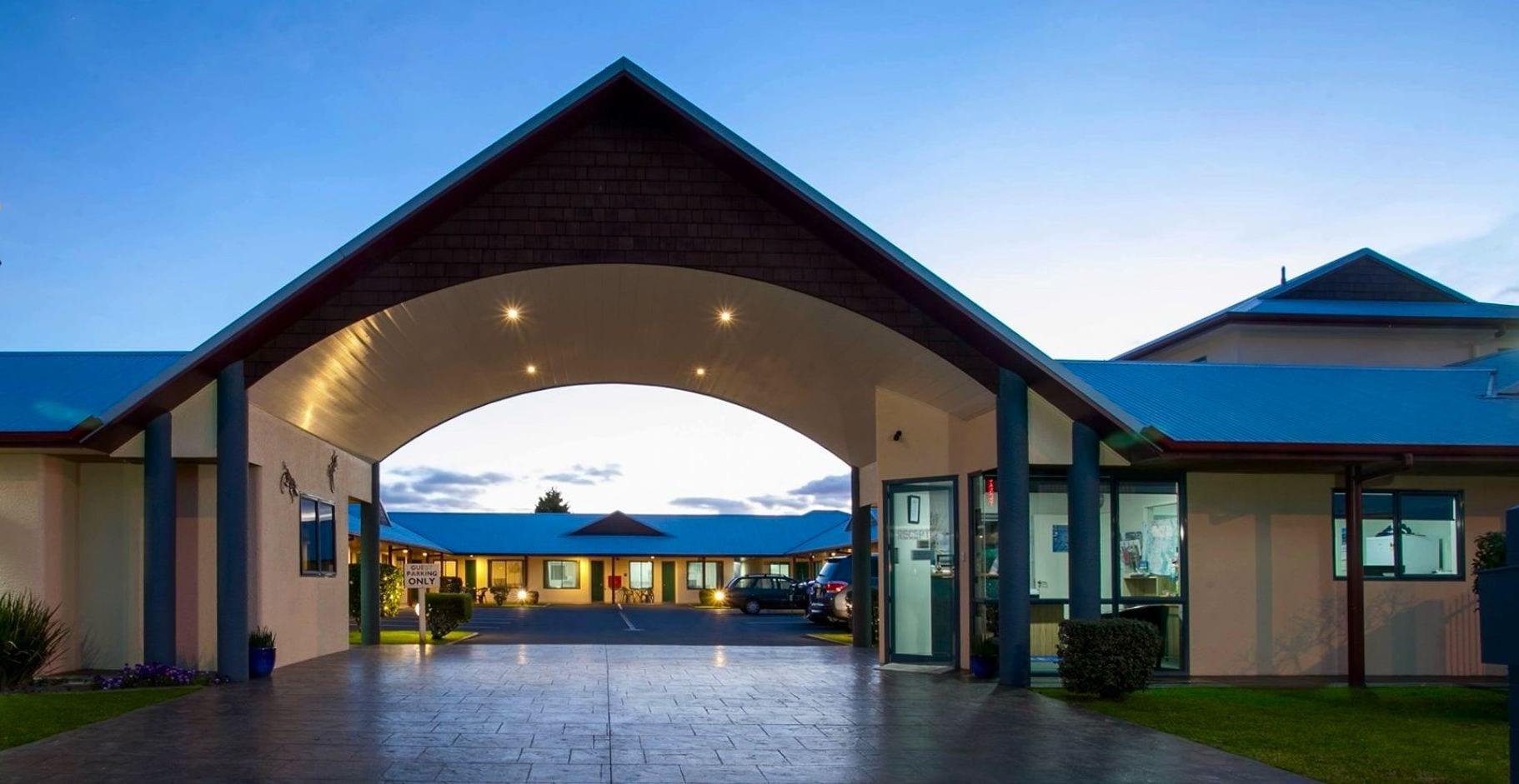 A modern hotel entrance with a covered walkway, a reception desk, and a parking area.