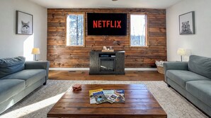 Uma sala de estar com lareira, uma televisão grande fixada em uma parede de madeira e dois sofás cinzentos.