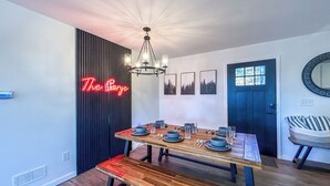 Uma área de refeições com mesa de madeira posta para quatro pessoas, um banco, um lustre e um letreiro de neon na parede.