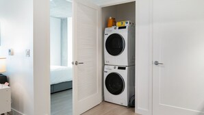 A laundry room with two stacked washing machines and a bed in the adjacent room. - Cozy 1-bedroom apartment in SF with AC, fitness room (San Francisco)