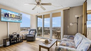 A spacious living room with a balcony, a ceiling fan, a flat-screen TV, and a view of the city. - Estero Beach and Tennis 1202 (Fort Myers Beach)