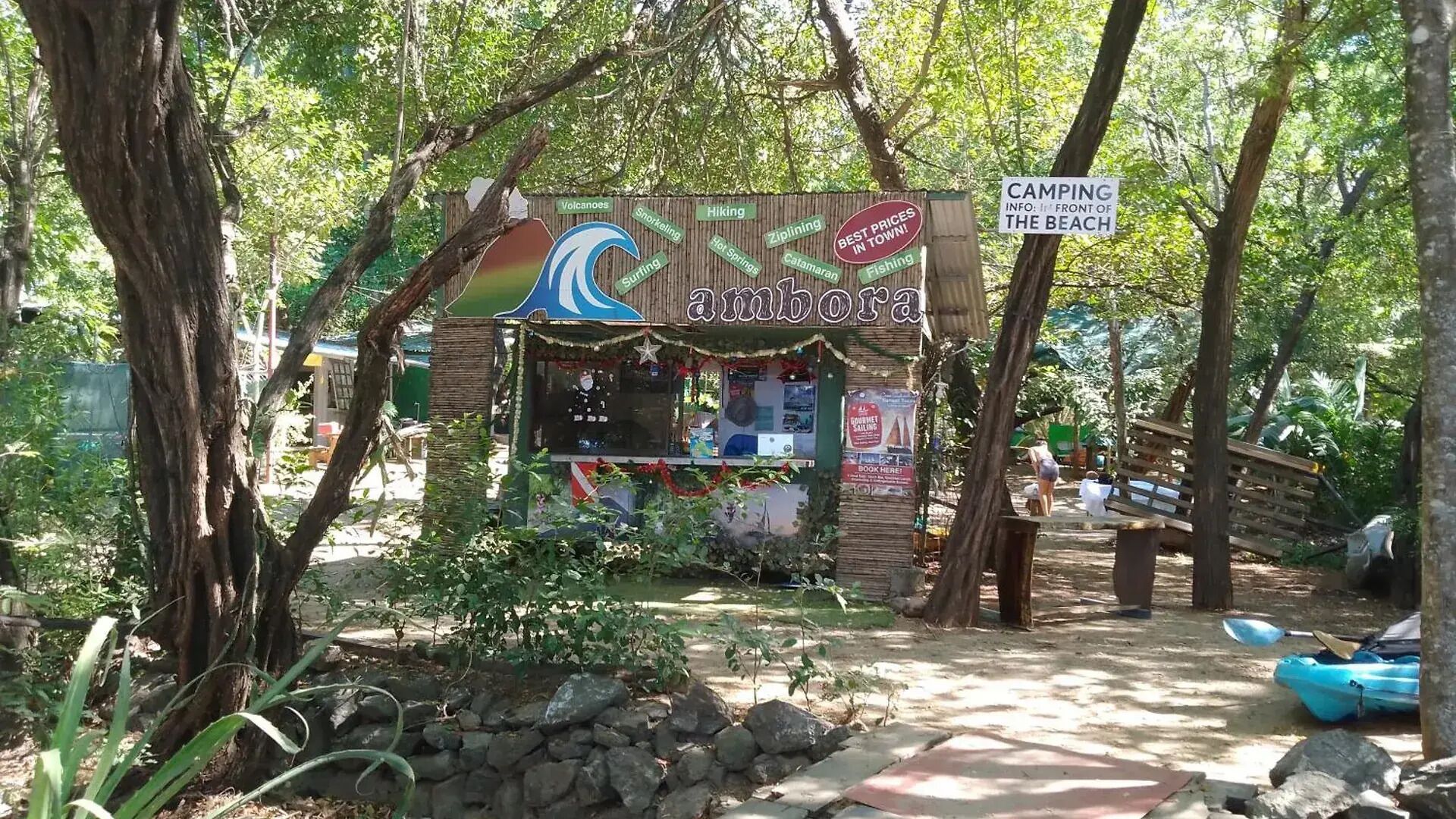 Un quiosco frente a la playa llamado 'Aniboga' con carteles anunciando camping, esnórquel, senderismo y tirolesa.