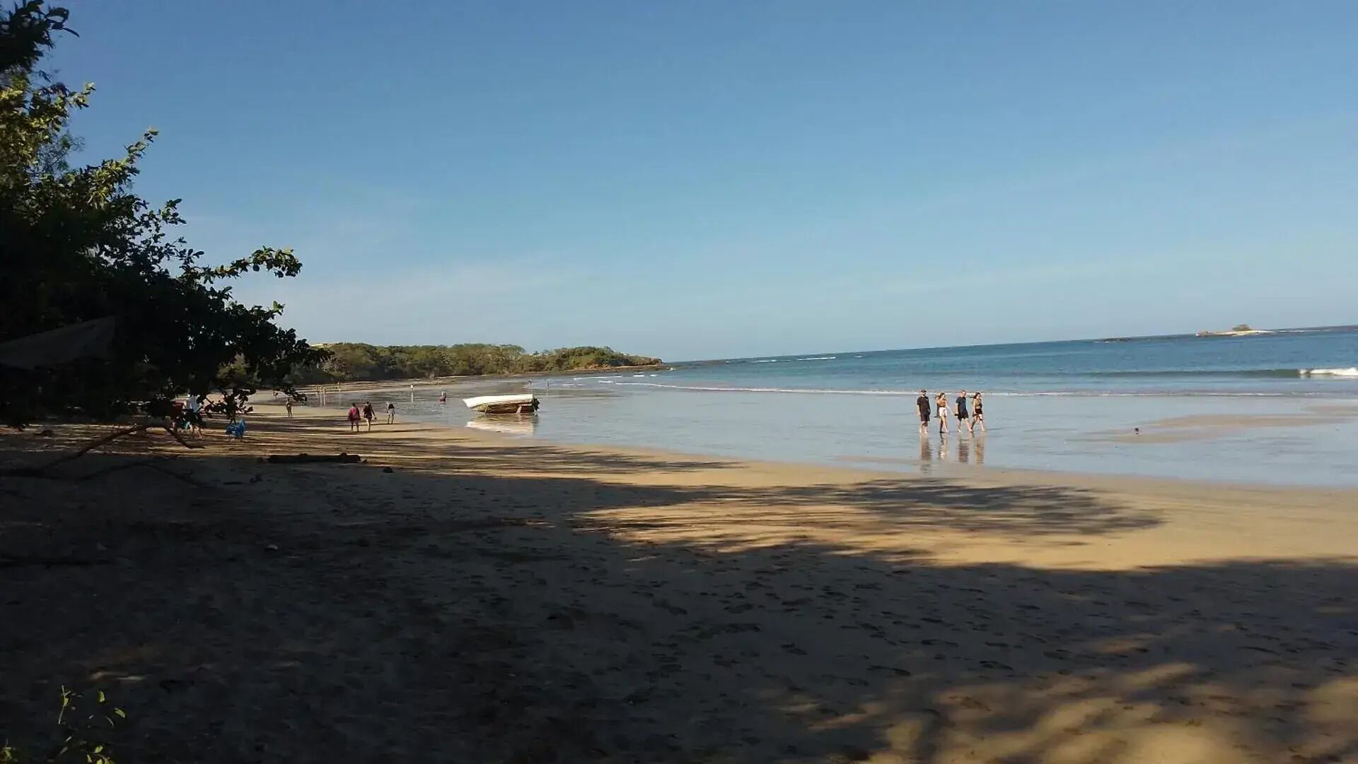 Una playa con gente caminando por la orilla, un barco en el agua y árboles al fondo.
