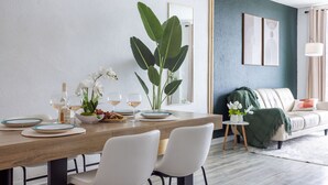 Un comedor moderno con una mesa de madera dispuesta para dos personas, rodeada de sillas blancas. Hay una planta verde grande sobre la mesa, junto con dos copas de vino y un cuenco con fruta.