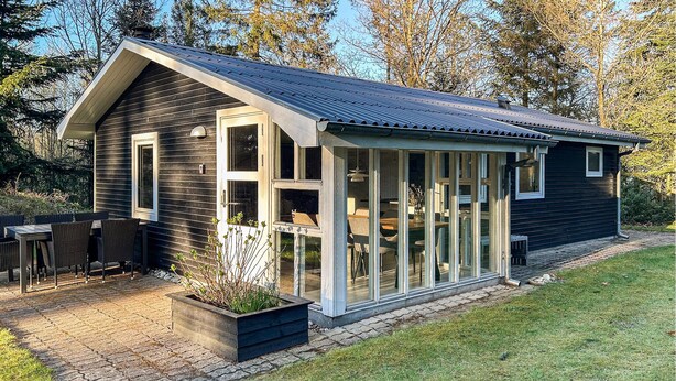 Et moderne hus med en stor glasudbygning, en terrasse med udendørs møbler og en velplejet have.