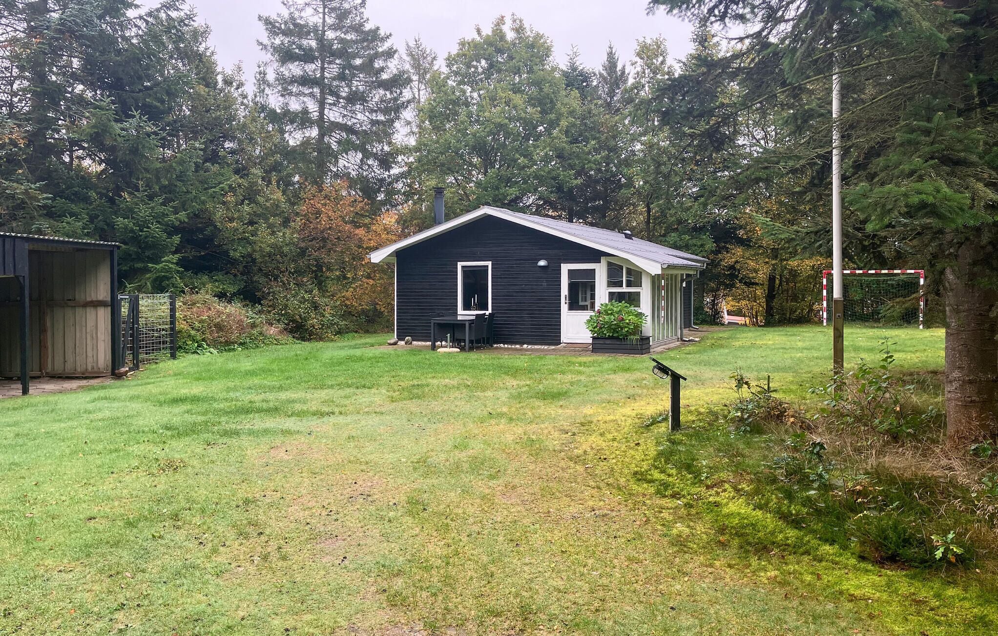 A small, single-story cabin with a dark exterior and white trim, surrounded by a well-maintained lawn and dense trees.