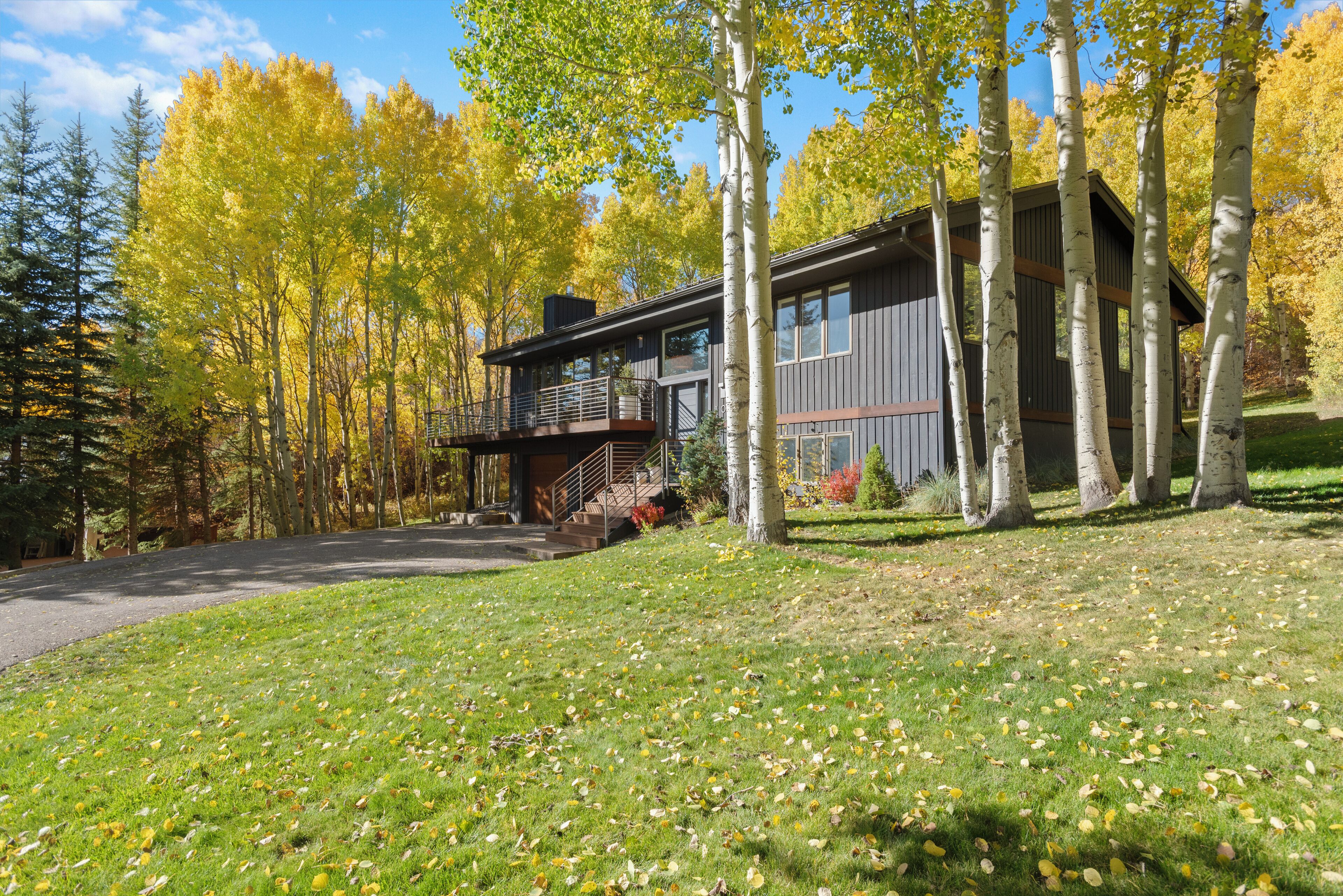 A modern two-story house with a balcony, surrounded by trees and a grassy lawn.