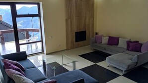 Un salon moderne avec un canapé gris, un canapé bleu et une table basse en verre. Un mur est recouvert de panneaux en bois et une grande fenêtre offre une vue sur les montagnes.