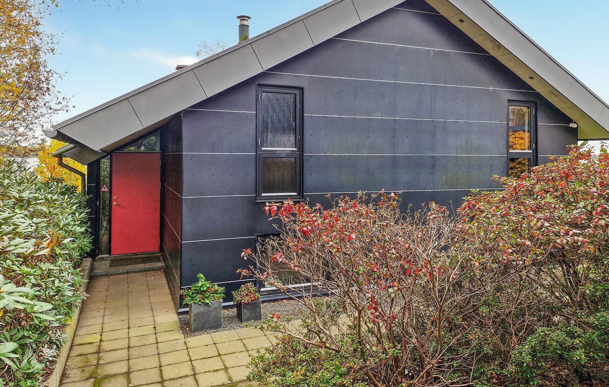 A modern building with a dark facade, a red door, and a small garden area with various plants.