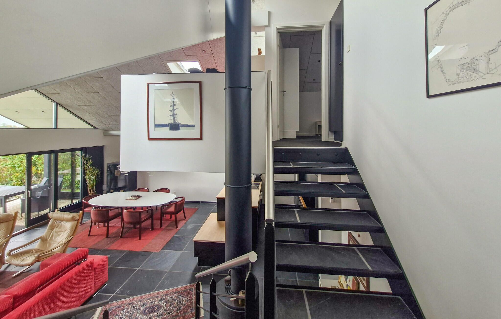A modern interior with a staircase, a dining area with a round table, and a large window overlooking greenery.