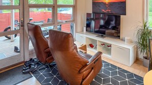 A modern living room with leather chairs, a flat-screen TV, and a large window with a view of a red fence and greenery. - Gorgeous home in Hovborg with WiFi (Hovborg)