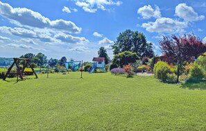 A playground with swings and slides, a small blue house, and a large tree.