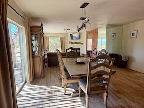 Un comedor amplio con una larga mesa de madera y sillas, un televisor montado en la pared y un gran ventanal con cortinas.