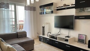 A modern living room with a large flat-screen TV, a sofa, and a coffee table. There are shelves above the TV and a small table with a vase and picture frame. - Luna Apartment - Sea View Private Apartment - AC - WiFi (Faro)
