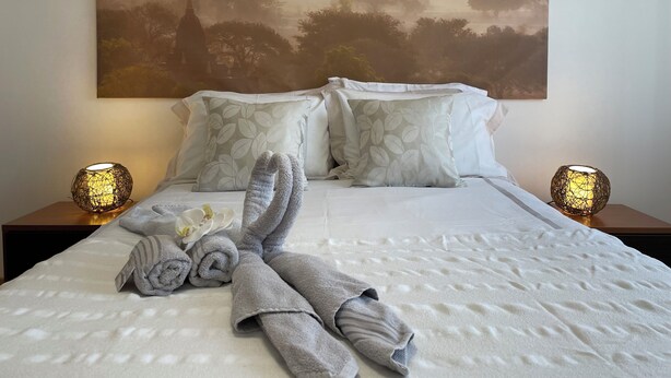 A neatly made bed with towels, a large wall painting, and two bedside lamps. - Luna Apartment - Sea View Private Apartment - AC - WiFi (Faro)