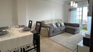 A modern living room with a dining area, featuring a grey sectional sofa, a dining table set with plates and glasses, and pendant lighting. - Luna Apartment - Sea View Private Apartment - AC - WiFi (Faro)