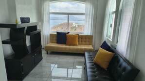 Ein Wohnzimmer mit einer Couch, einem schwarzen Sofa, einer TV-Bank und Blick auf ein Gewässer durch ein großes Fenster.