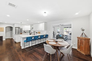 A modern kitchen with white cabinets, a central island with blue bar stools, a dining area with a round table and grey chairs, and a wooden cabinet with a vintage lamp.