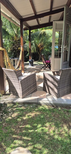Een overdekt terras met rieten meubilair, een hangmat en een deur naar een binnenruimte.