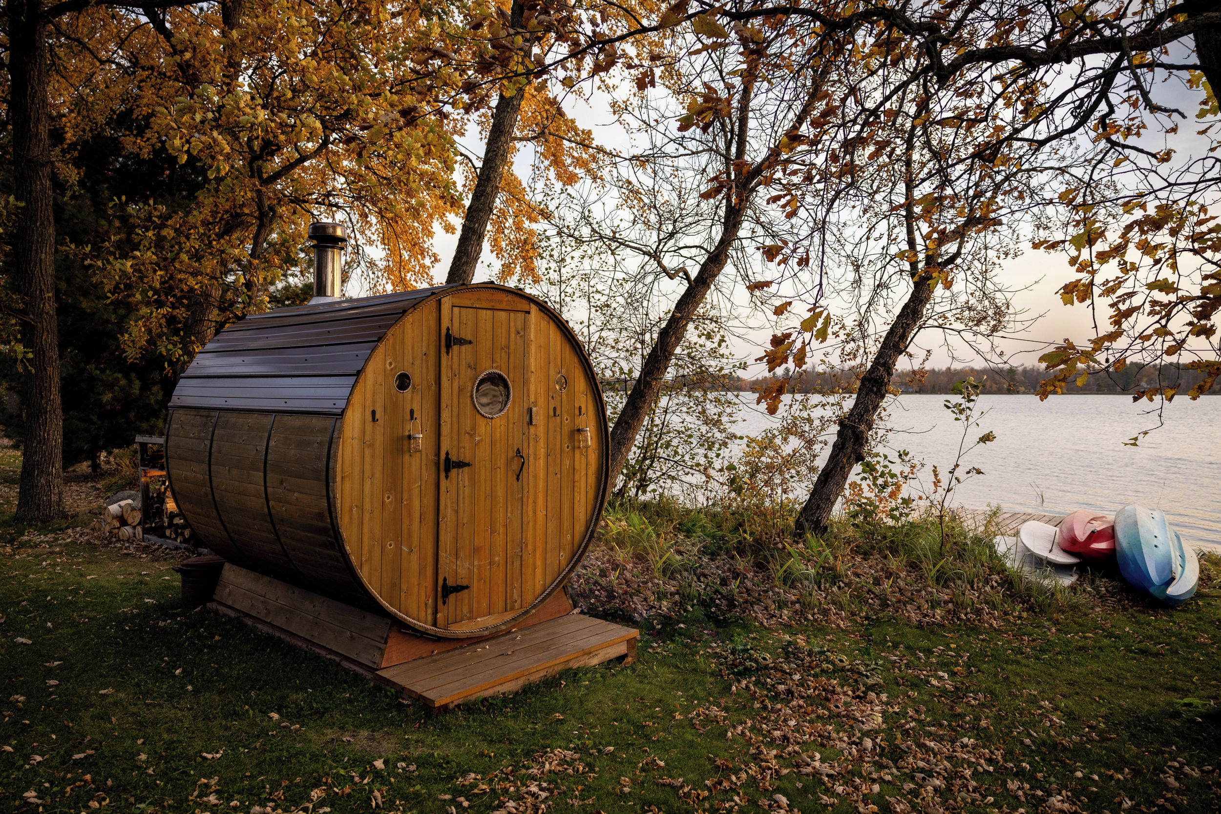 Une structure de sauna en bois, dotée d’une cheminée, située au bord d’un lac, avec des bateaux à proximité.
