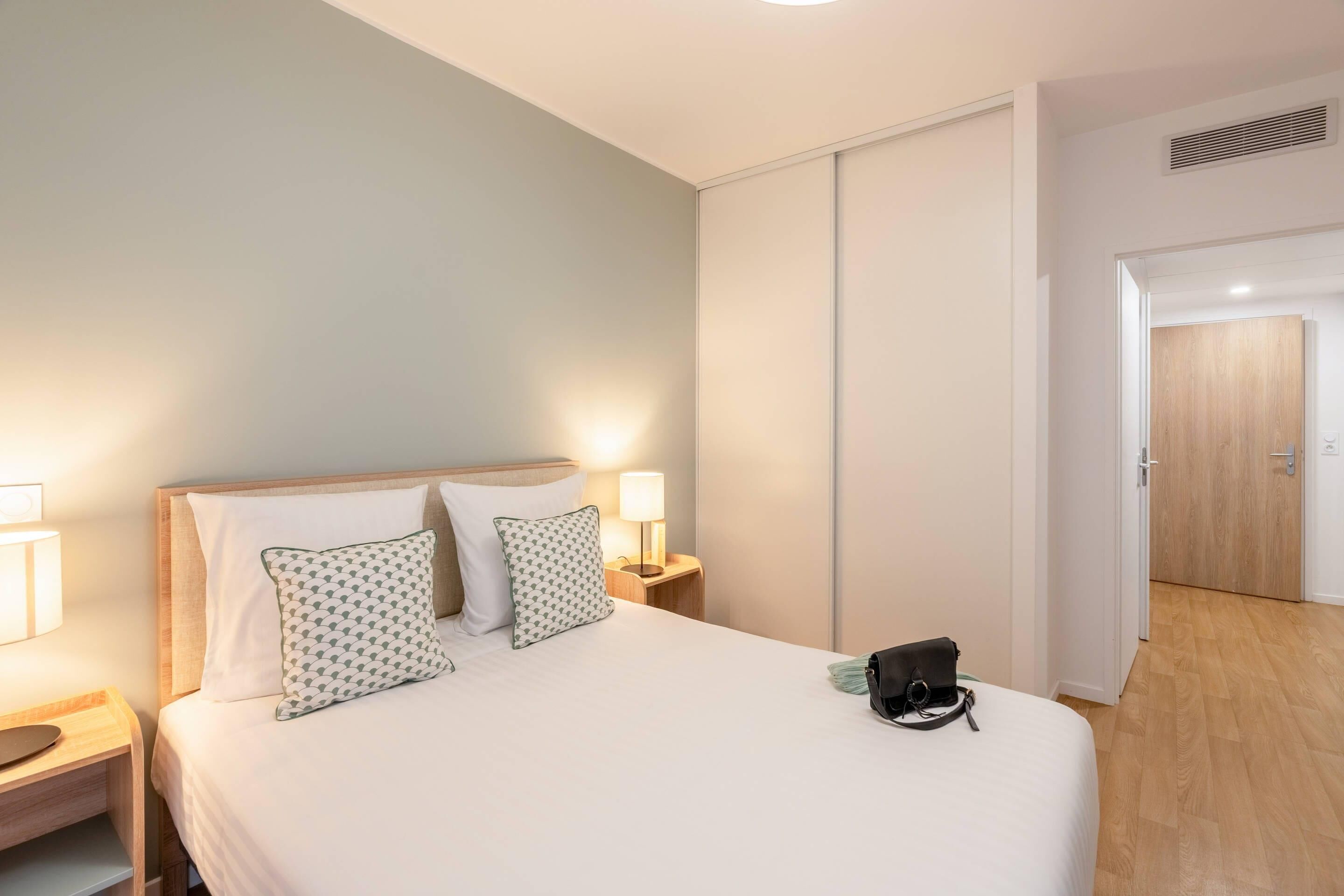 A hotel room with a bed, bedside lamps, a nightstand, a door, and a wooden floor.