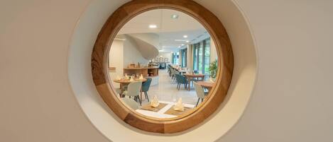 Una ventana circular con marco de madera que muestra un moderno comedor con mesas y sillas.