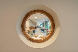 Una ventana circular con marco de madera que muestra un moderno comedor con mesas y sillas.
