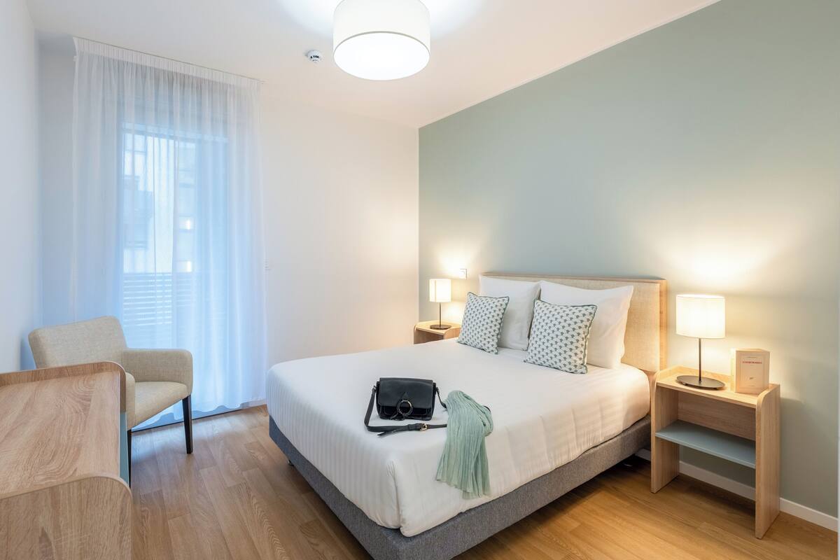 A hotel room with a bed, bedside tables, a chair, and a window with curtains.