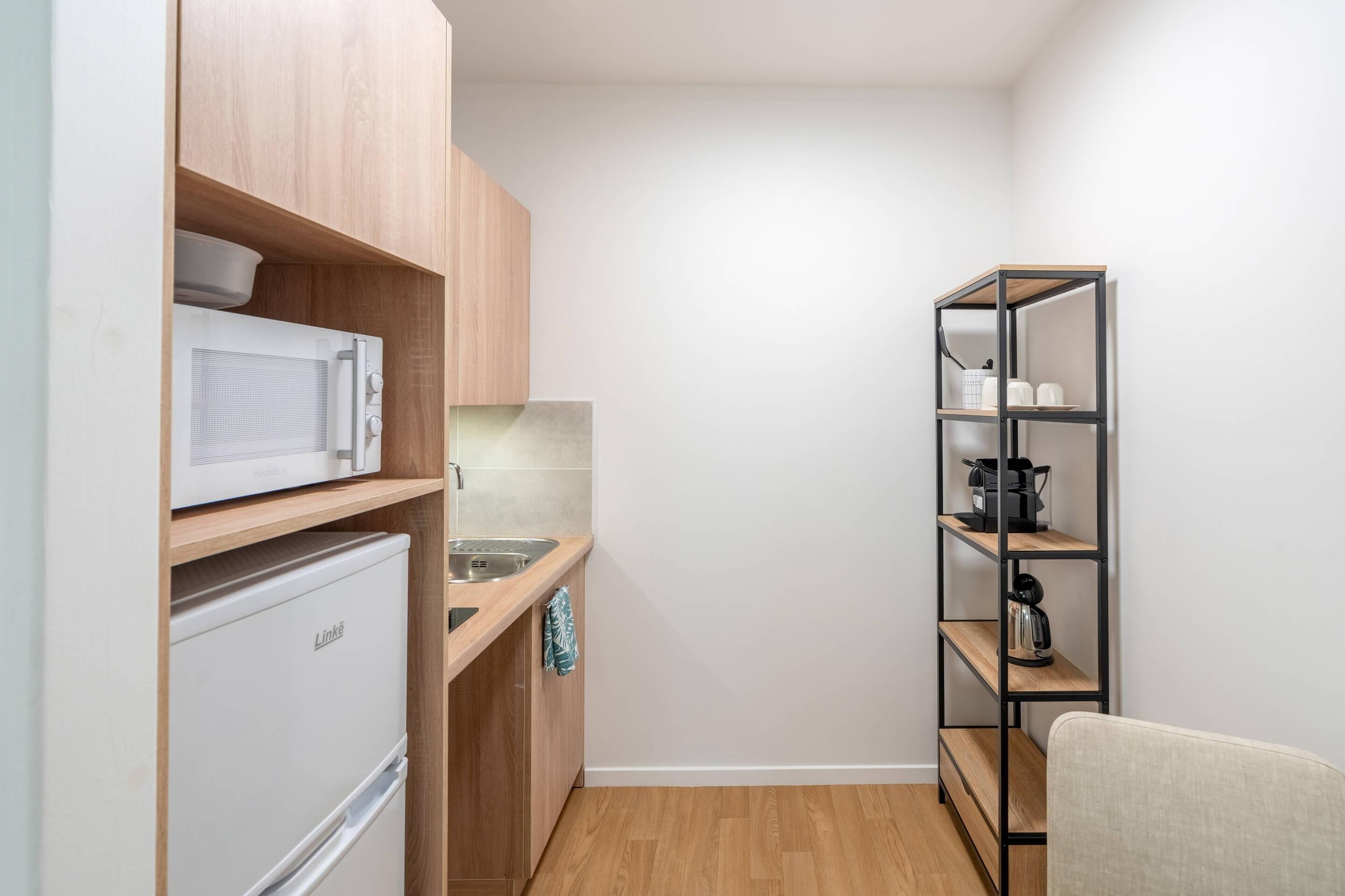 A compact kitchen with wooden cabinets, a microwave, a Linke refrigerator, a sink, and a metal shelving unit.