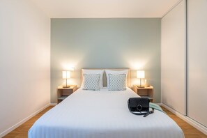 A neatly made bed with a white bedspread and two patterned pillows, flanked by bedside tables with lamps, in a room with light-colored walls and wooden flooring. - Quality Aparthotel Nancy Zenith (Laxou)