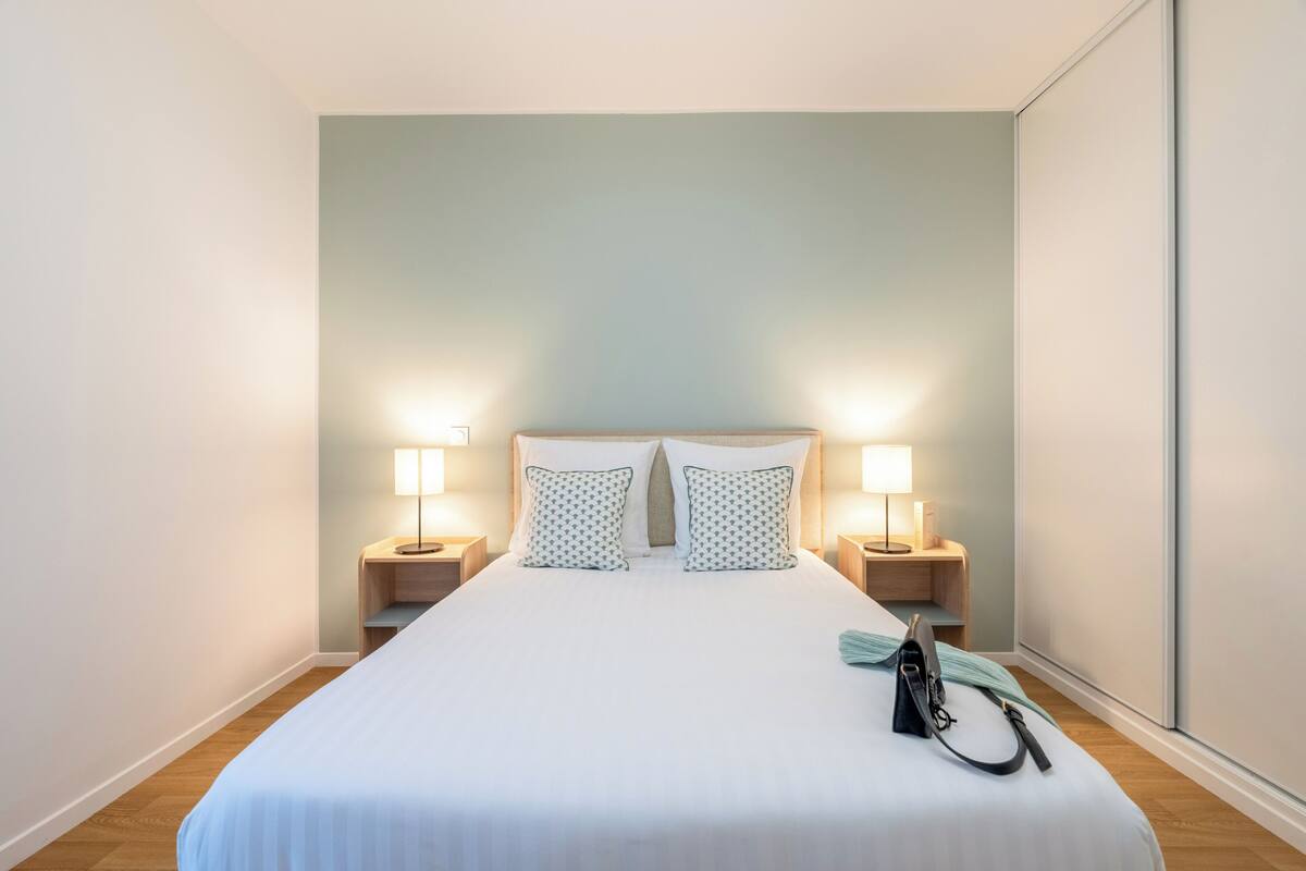 A neatly made bed with white bedding and patterned pillows, two bedside lamps, a small wooden nightstand, and a black bag on the bed.