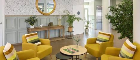 A lobby with yellow armchairs, a round table with a magazine, a mirror, and a reception desk with plants.