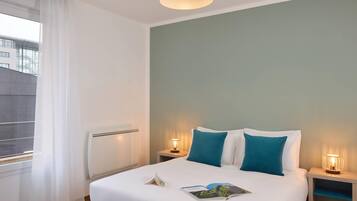 Habitación de hotel con cama, dos mesitas de noche con lámparas, ventana con cortinas translúcidas y vista a edificios.