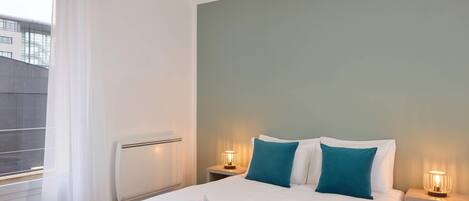 A hotel room with a bed, two bedside tables with lamps, a window with sheer curtains, and a view of buildings outside.