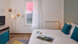 A hotel room with a bed, a TV, a desk, a floor lamp, and a window with a curtain. - Quality Aparthotel Valenciennes (Valenciennes)