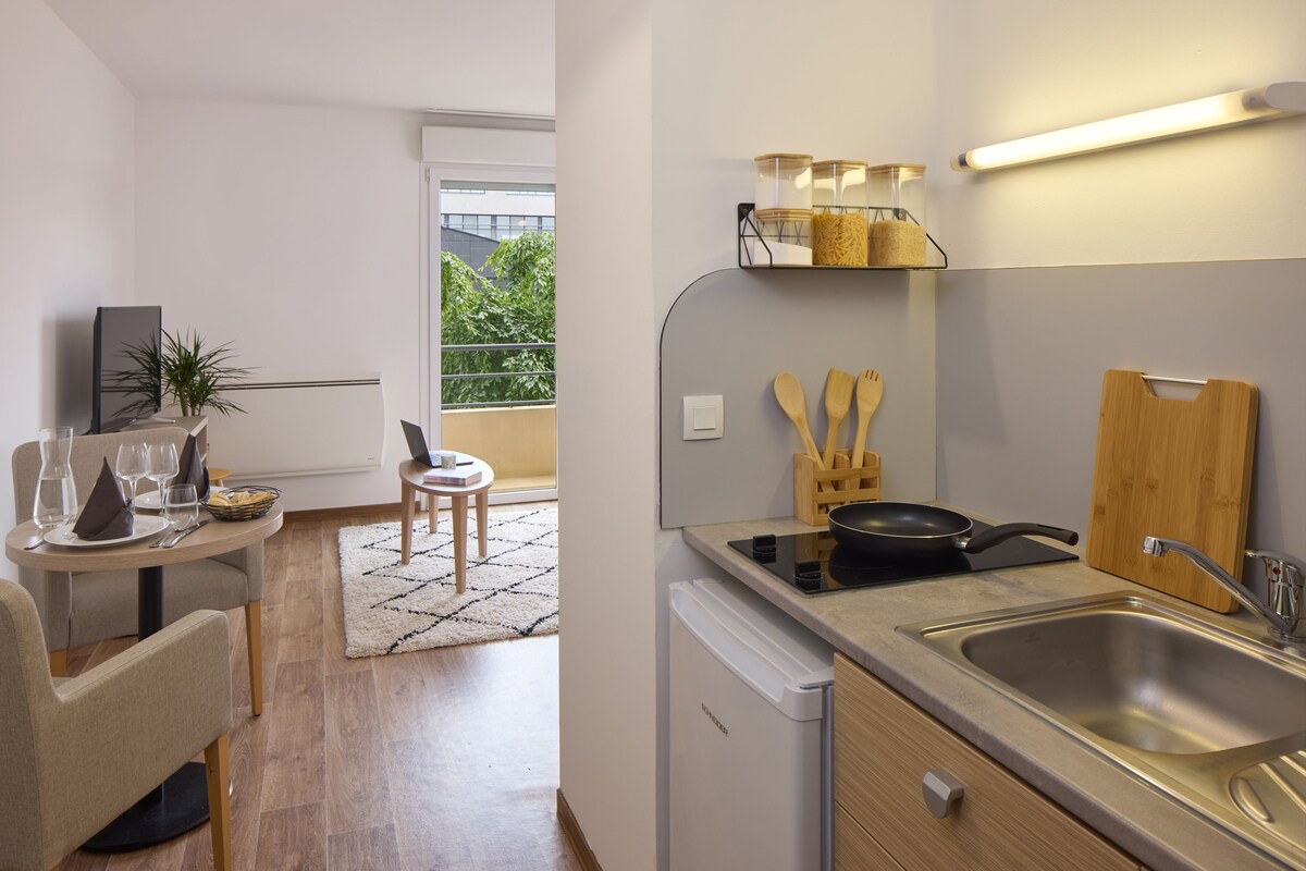 Een moderne keuken met een vaatwasser, kookplaat, spoelbak en houten keukengerei. Een eethoek met een tafel en stoelen, en uitzicht op groen.