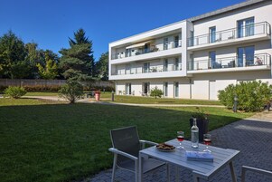 Un edificio de apartamentos moderno con varios balcones, un césped cuidado y un patio con mobiliario exterior.