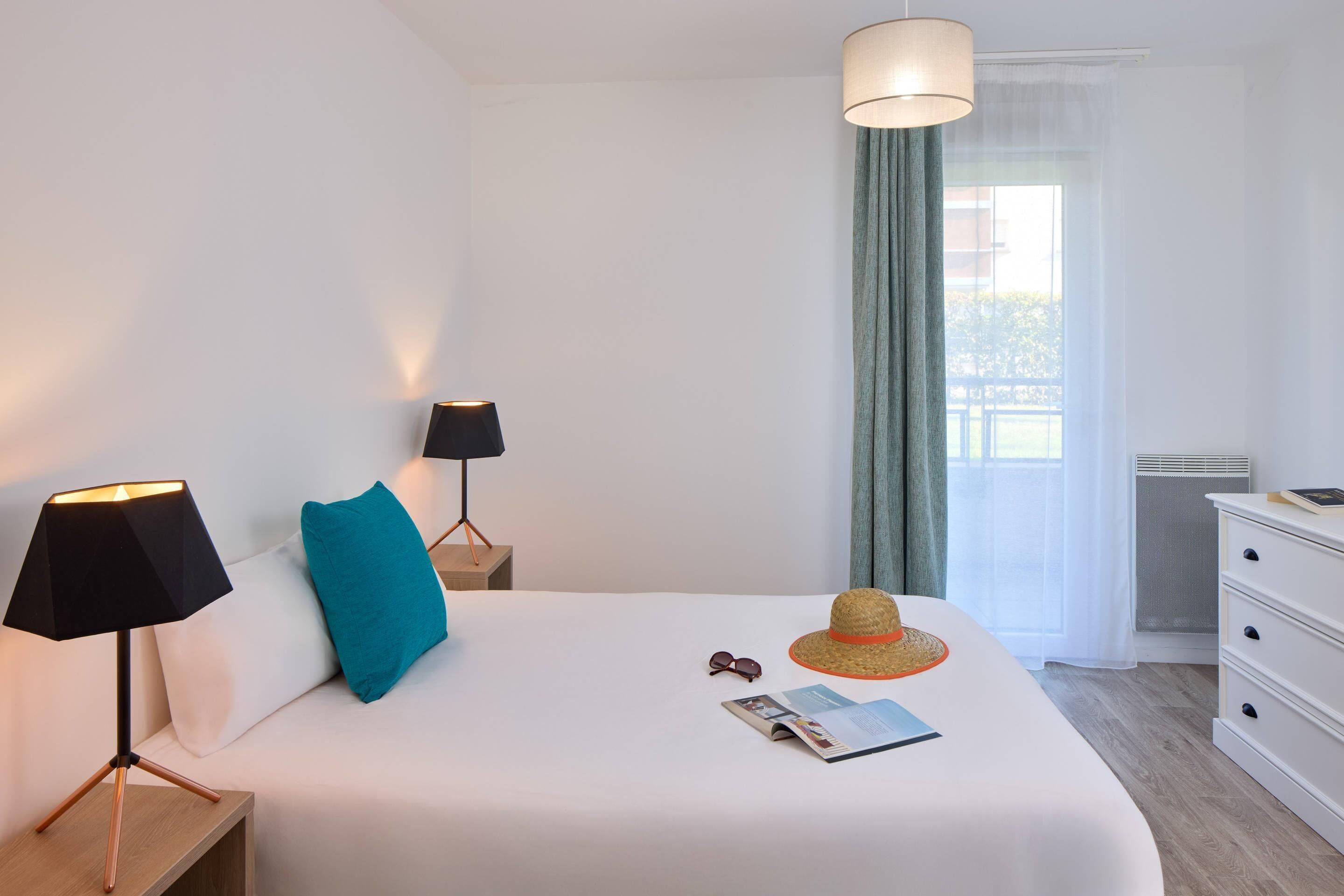 A neatly made bed with a white comforter, a blue pillow, a straw hat, and a pair of sunglasses on the bed. There is a black lamp on a wooden nightstand beside the bed.