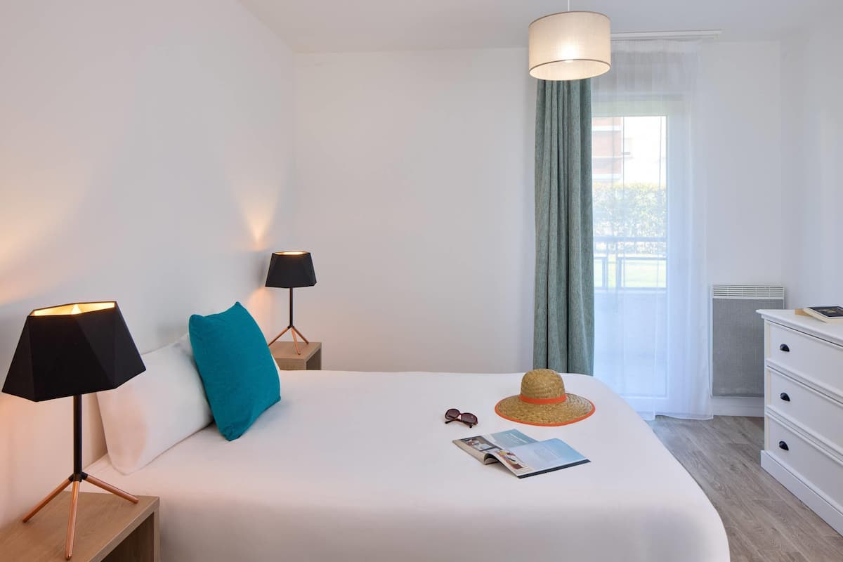 A neatly made bed with a white comforter, a blue pillow, a straw hat, and a pair of sunglasses on the bed. There is a black lamp on a wooden nightstand beside the bed.