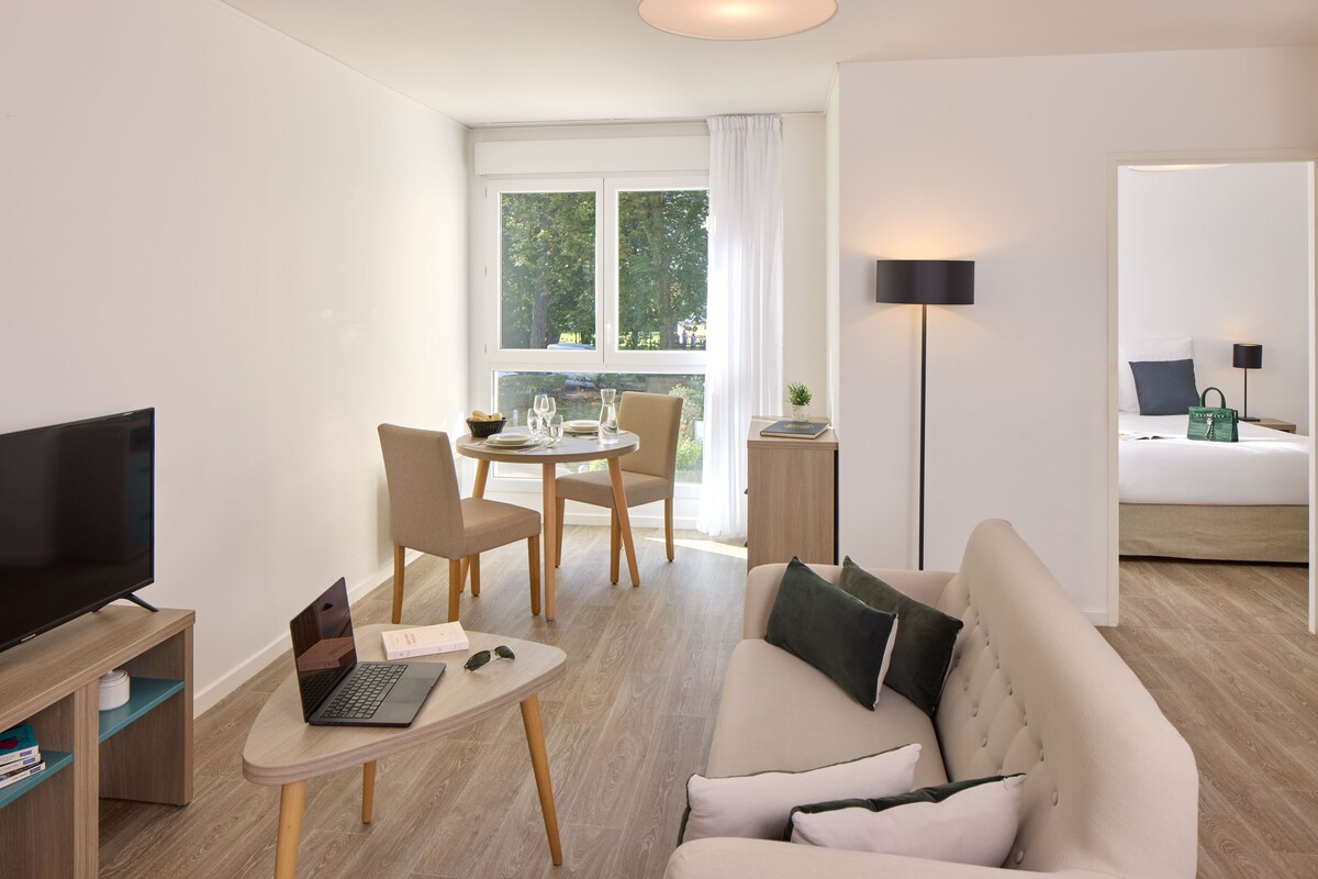 A modern hotel room with a flat-screen TV, a laptop on a small table, a sofa, a dining area with chairs, and a bed with a nightstand.