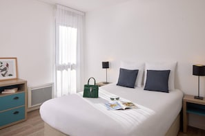 A neatly made bed with pillows, a bedside table with a lamp, a framed picture, and a window with curtains. - Quality Aparthotel St-Quentin en Yvelines (Montigny-le-Bretonneux)