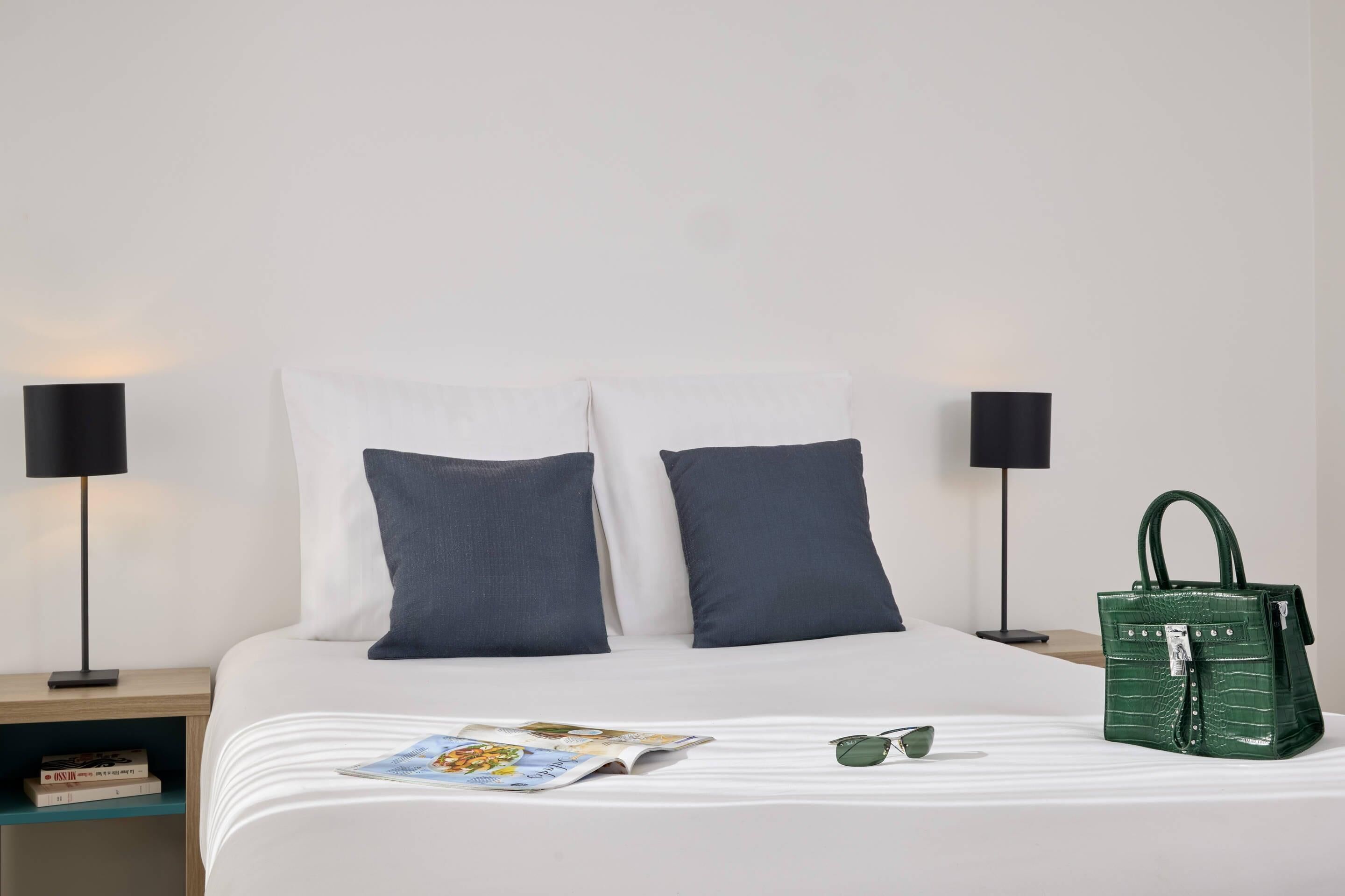 A neatly made bed with two pillows, a green handbag, and a magazine on the bed.
