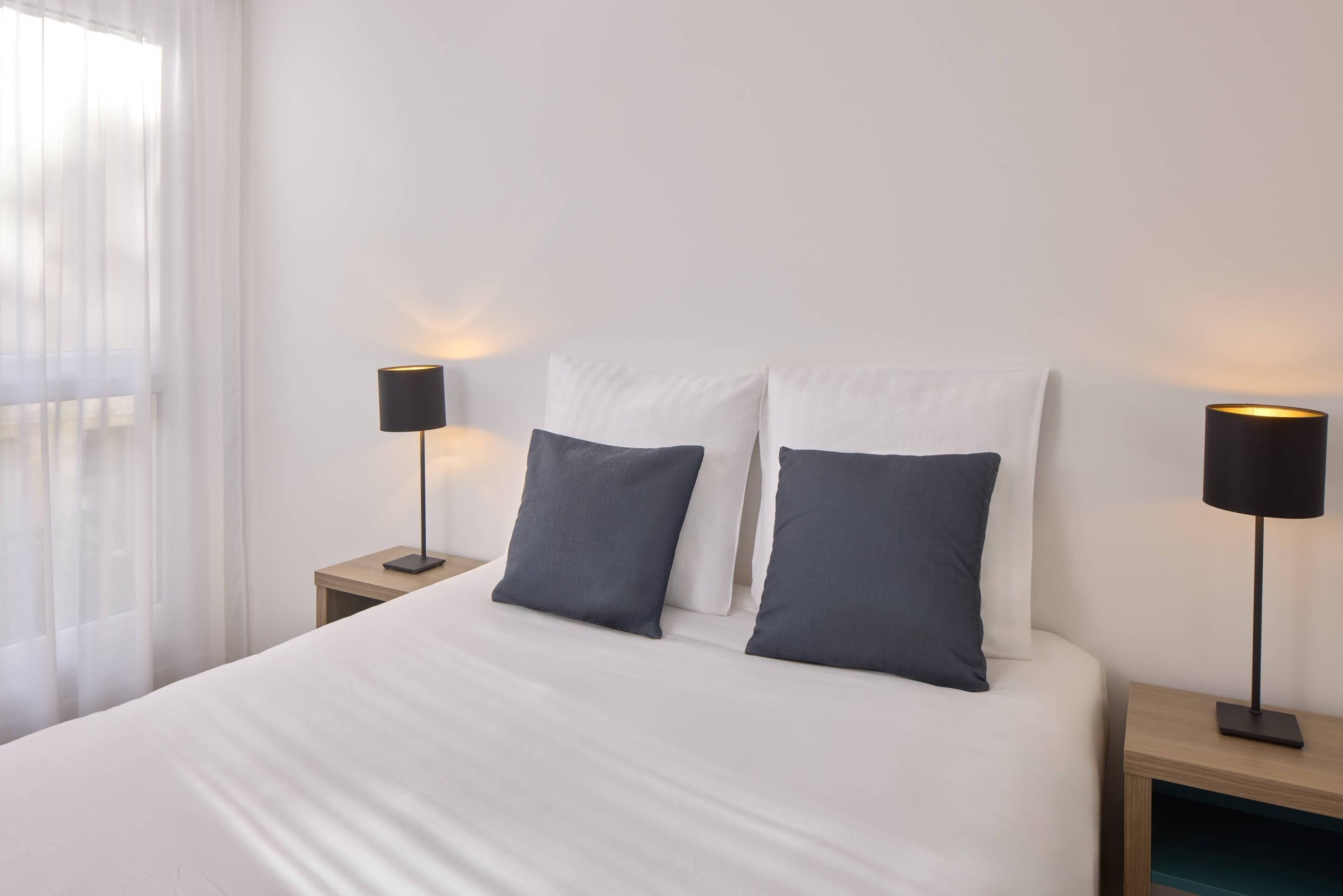 A neatly made bed with white bedding and two pillows, flanked by two bedside tables with black lamps.