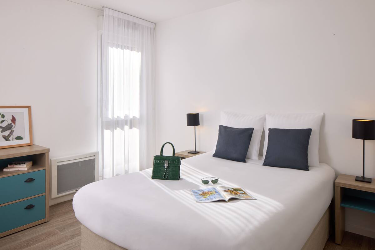 Une chambre d’hôtel bien rangée, avec un lit, des tables de chevet, une commode, un tableau encadré, une fenêtre avec des rideaux et un radiateur.
