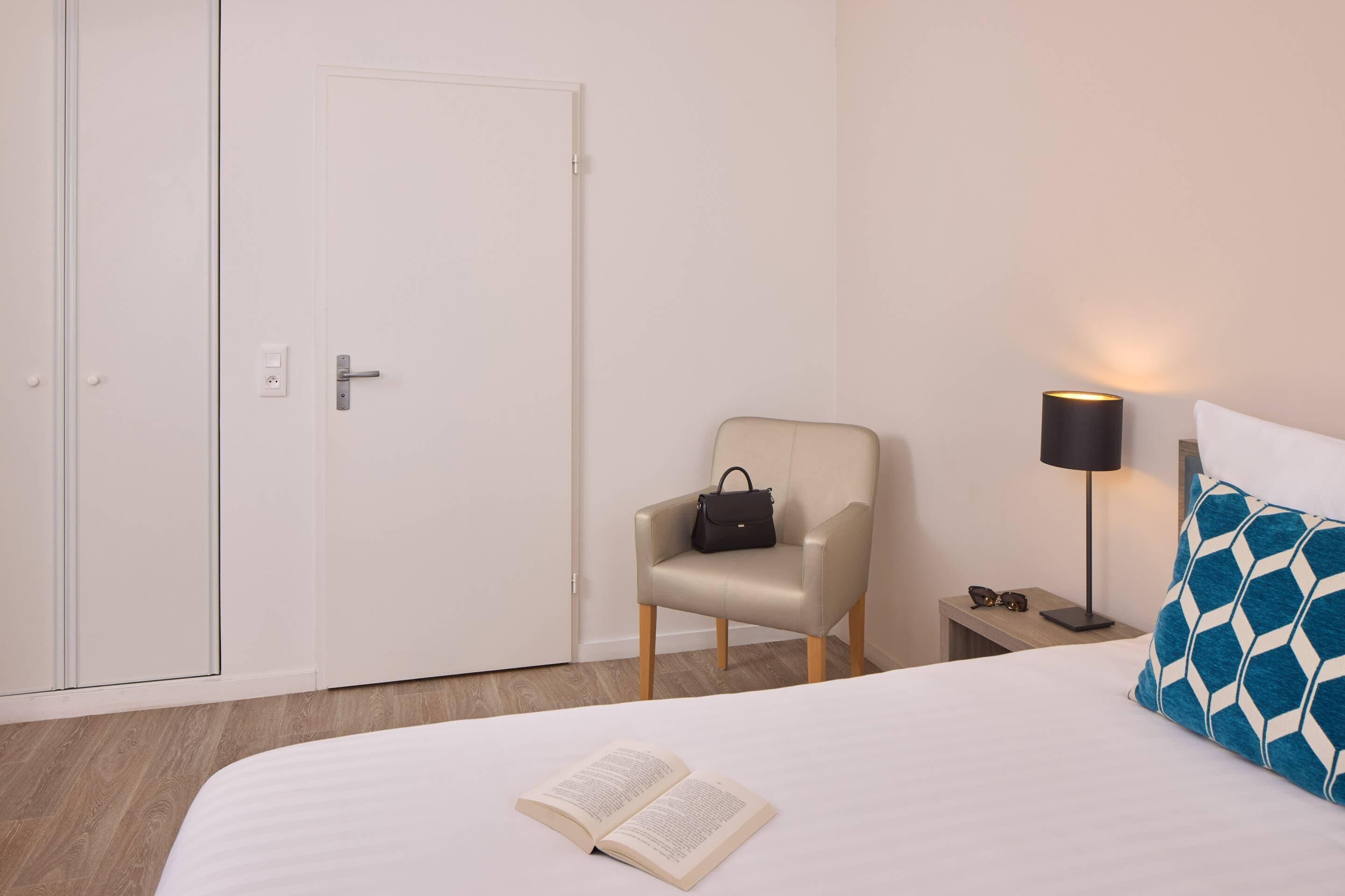 A hotel room with a bed, a chair, a bedside table with a lamp, and a wardrobe.