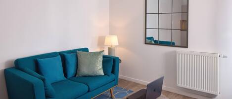 A teal sofa with a lamp and a laptop on a small table, a mirror on the wall, and a radiator.
