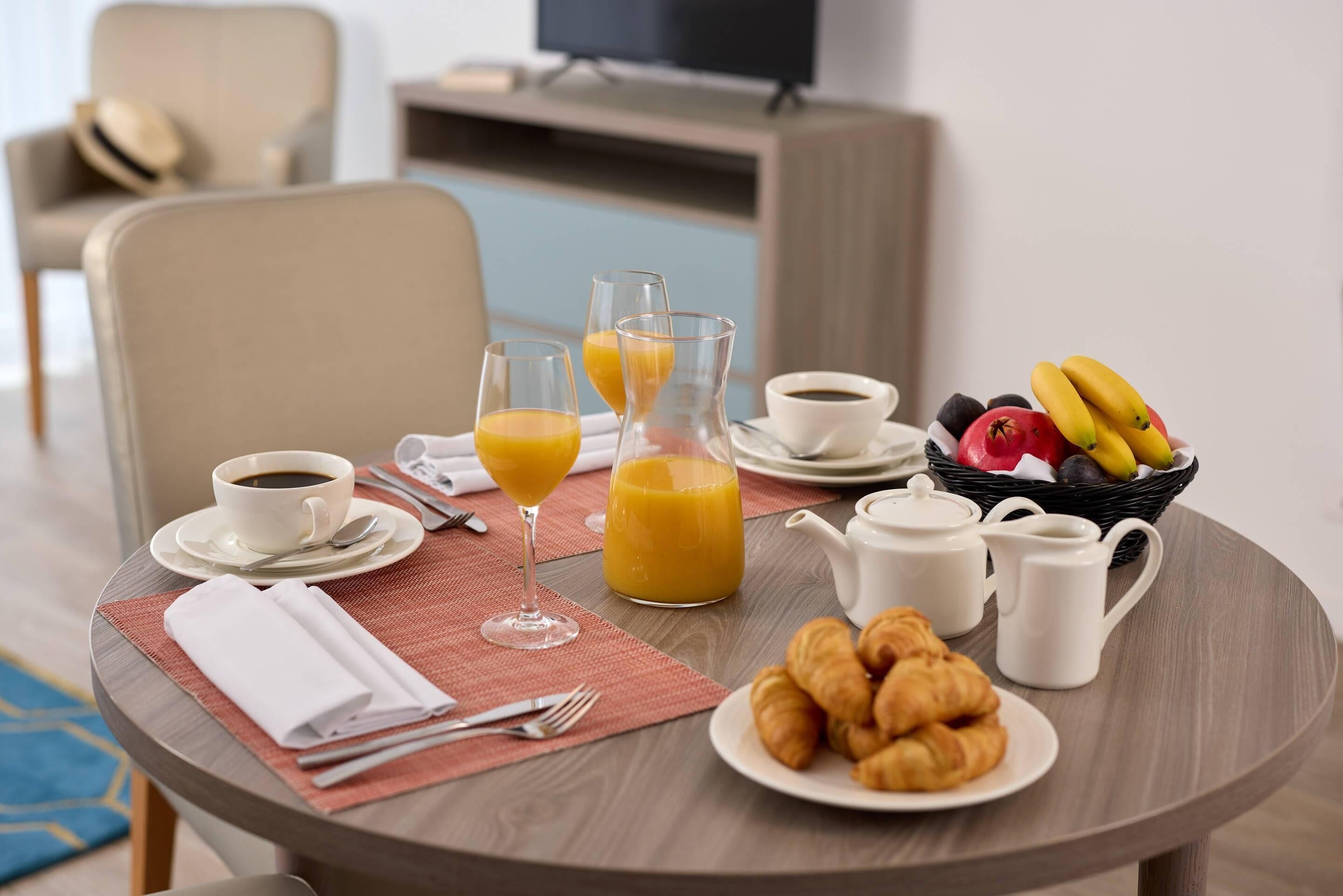 A breakfast table with croissants, juice, coffee, and a fruit basket.