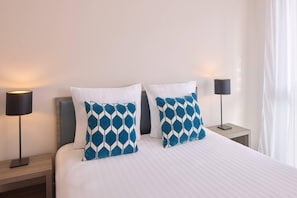 A neatly made bed with white bedding and two blue patterned pillows. There are two bedside tables, each with a black lamp. The room has a clean, minimalist design with light-colored walls. - Quality Aparthotel Morangis Orly (Morangis)